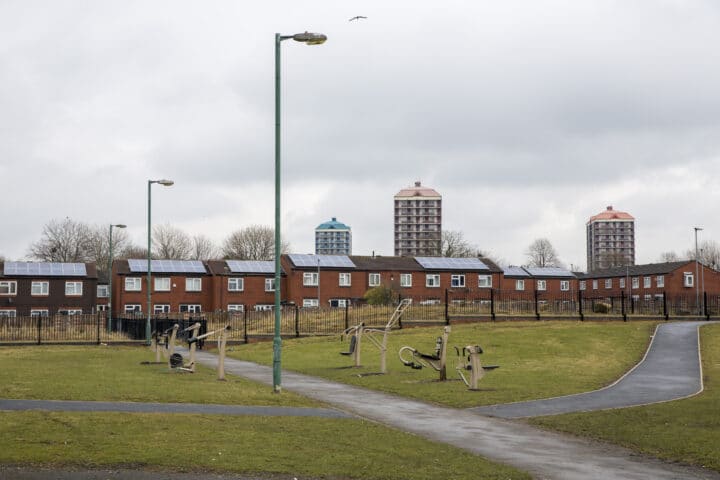A row of new-build houses with sloar panels on the roof. In teh distance are high rise flats. In the forground is a park with exercise equipment.