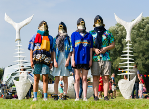 Four people standing on a grassy field wearing fish head masks and colourful holiday-style clothing, including Hawaiian shirts, shorts, and scarves. Behind them, there are large fishbone sculptures with tail fins, set up in an outdoor festival setting with people, tents, and decorations in the background. 