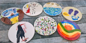 This image displays seven hand-painted ceramic plaques arranged on a wooden surface. Each plaque features a unique design, including a coastal scene with cliffs and a boat, a stick-figure drawing with the words "Those Were the Days," an abstract splatter painting, a black bird labelled "Cornish Chough," a vibrant rainbow landscape with a sun, a seaside water scene, and a collection of summer-themed items such as flip-flops, a sun hat, and a watering can.