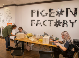 Three people working around a table. The table is full of craft material, and the stylised decal on the wall behind the table says 'Pigeon Factory'