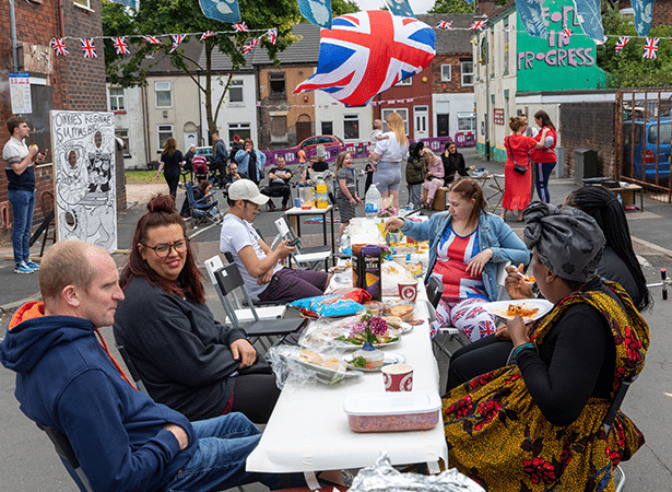 A lively street party with people gathered around a long table covered in food, drinks, and containers. The setting is an urban residential area with terraced houses in the background, decorated with Union Jack bunting and flags. A large inflatable Union Jack balloon floats above the scene. People of diverse backgrounds are engaged in conversation, eating, and enjoying the communal atmosphere.