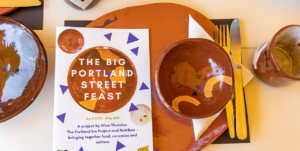 A close-up photo of a flyer titled "The Big Portland Street Feast" with the dates April 2020 - May 2021, by Alice Thatcher. The flyer is surrounded by handmade ceramic plates, bowls, and cutlery on a light-colored surface.