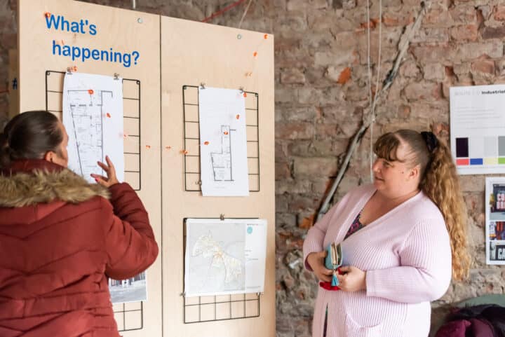 Woman in red coat and woman in pink cardigan look at a floorplan pinned to a board.