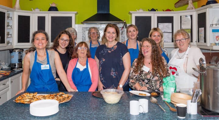 A group of women of mixed ages stand in a kitchen at a community event smiling at the camera. Some women are wearing blue aprons.