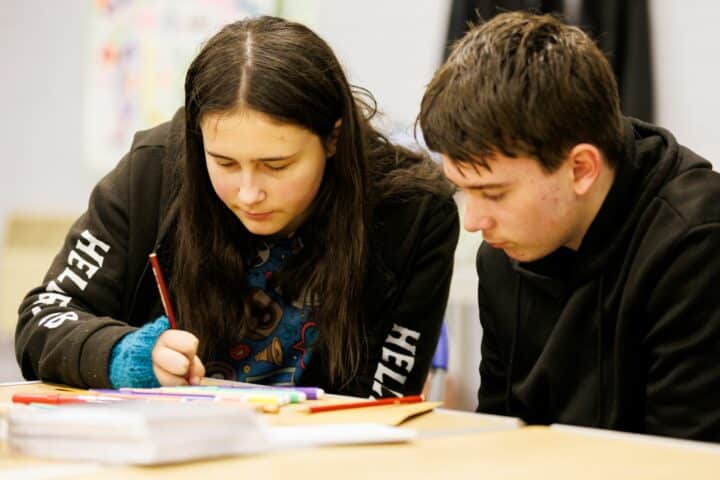 Two young people lean over some paper and draw in it on it using coloured pens