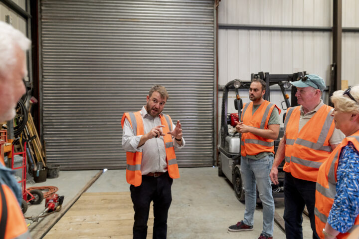 Five people in a warehouse wearing orange high vis jackets. One is talking to the group.