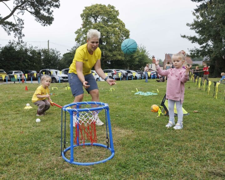 Young children take part in an outdoor basketball activity with an instructor wearing a yellow tshirt