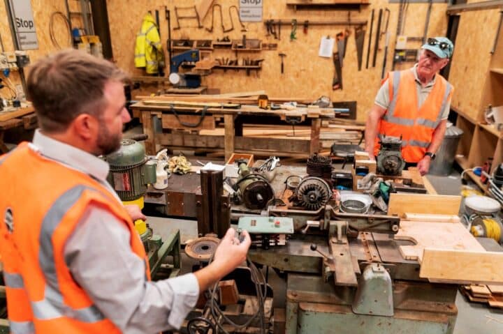 Two men in a workshop with tools on the wall, looking at some mechanical equipment