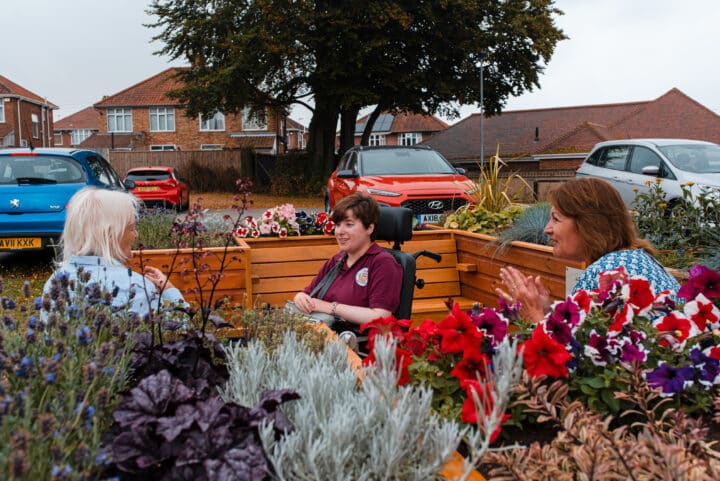 Three seated women are chatting, surrounded by colourful flowers . One woman is in a wheelchair, and there are parked cars and houses visible in the background.