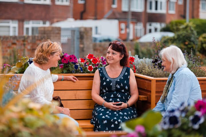 Three women sit on benches with flowers around them chatting