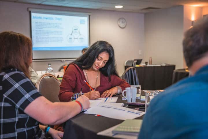 Image of a woman in a deep red blouse seated at a table, writing on paper with an orange pencil. Two other people are seated around her in a conference hall setting, with paper, mugs, and cups on the table. A presentation slide is visible in the background.