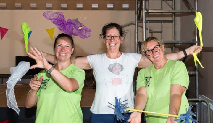 Three cheerful individuals, two wearing bright green t-shirts, are smiling energetically and holding colourful props, including lightweight netting and playful sticks with attachments. The setting is decorated with colourful bunting. The person in the centre is wearing a white t-shirt featuring a graphic print of Princess Leia and the words 