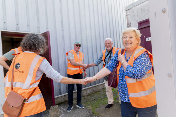 A group of individuals wearing orange high-visibility vests, smiling and shaking hands in an outdoor setting. One person is prominently in the foreground smiling towards the camera.