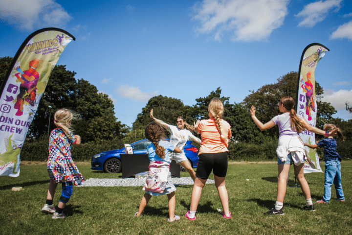 A group of children participating in an outdoor dance activity led by an instructor in a park-like setting. Two large banners reading 