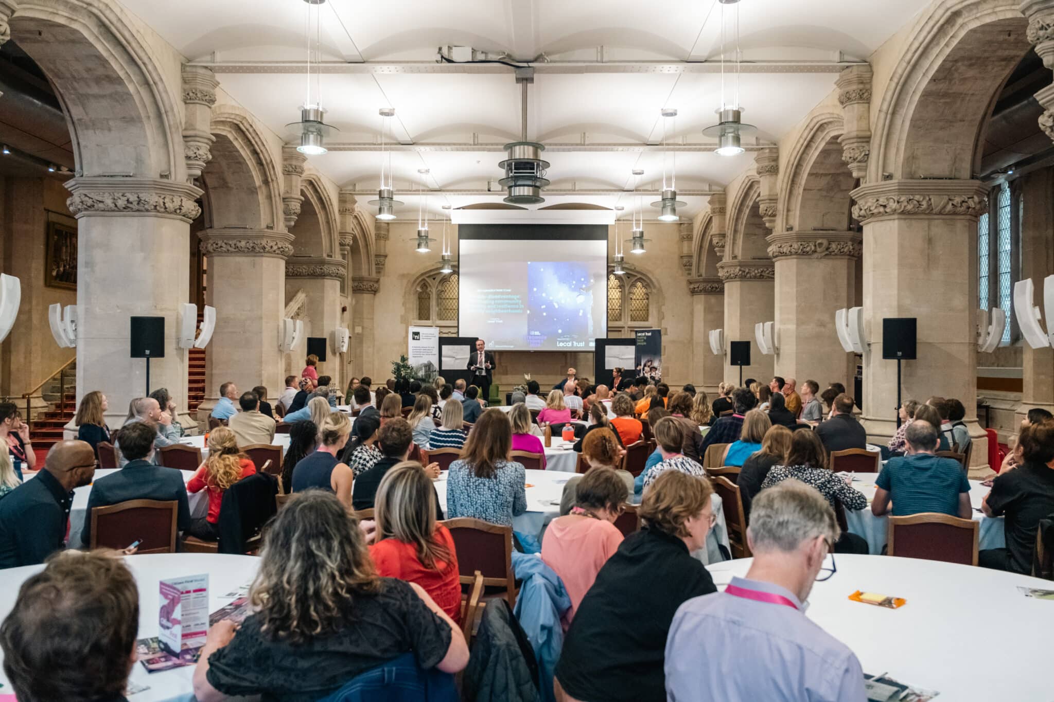 A large gathering of people attending a conference in a grand hall with high, arched ceilings and stone columns. The audience is seated at round tables, facing a stage where a speaker is presenting in front of a large screen.