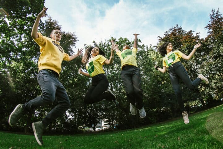 Four young people in an outdoor setting jump in the air with arms outstretched. They are wearing matching yellow t-shirts.