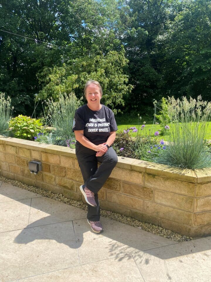 Woman wearing a black t-shirt and trousers is smiling as she leans on a low wooden wall. Behind her, there is a lush green garden with various plants and flowers, and a backdrop of tall trees.