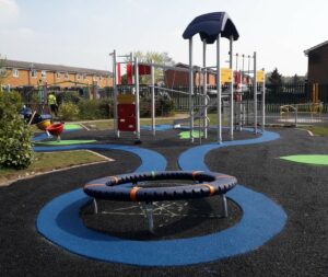 An image of Diamond Park - a modern playground with various play structures, including a climbing frame with a slide, a rope bridge, and a circular rope swing. The flooring is made of colourful rubber safety surfacing in black, blue, and green patterns. There are benches and greenery surrounding the play area. 