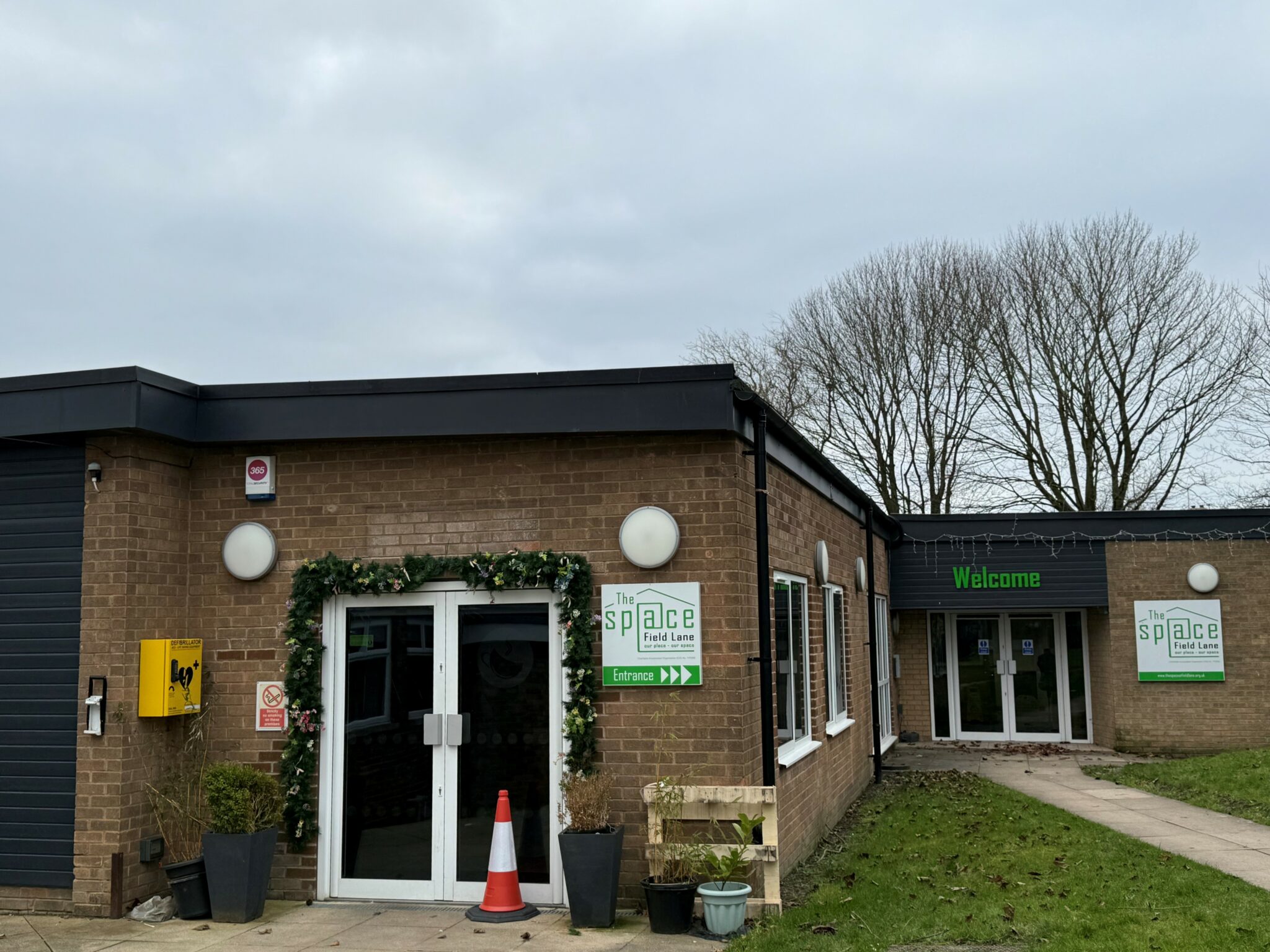 A modern, single-story brick building with two entrances, labeled "The Space @ Field Lane." One entrance has glass double doors decorated with greenery, and the other has a "Welcome" sign above. The sky is overcast, and leafless trees stand in the background.