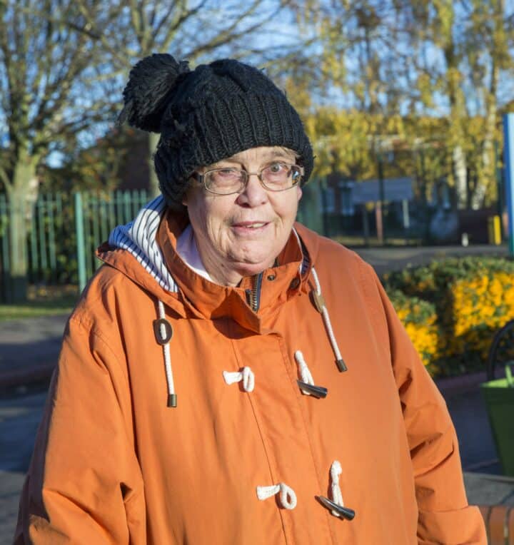 Yvonne Griggs wearing a black knitted beanie with a pom-pom and glasses is outdoors on a sunny day. She is dressed in an orange duffle coat with white rope fastenings and a striped hood lining. The background features trees with autumn foliage, a green fence, and a landscaped area with yellow flowers.