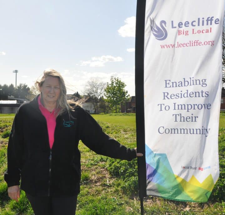 Donns Inwood stands outdoors on a grassy area next to a vertical banner. She is wearing a black fleece jacket over a pink polo shirt and has long blonde hair. The banner beside her reads: 