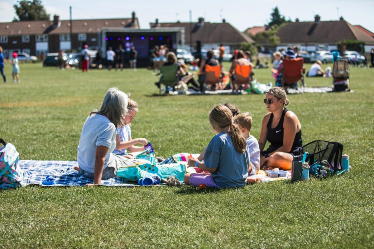 A group of adults and children sit on a picnic blanket enjoying snacks on a sunny field, with others seated nearby. In the background, a small outdoor stage hosts a live performance, and rows of houses line the far edge of the green space.