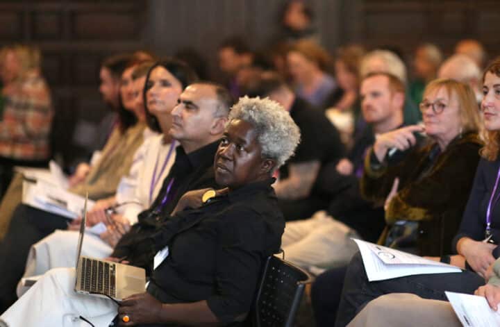 Rows of conference attendees face forwards as they listen to a speaker.