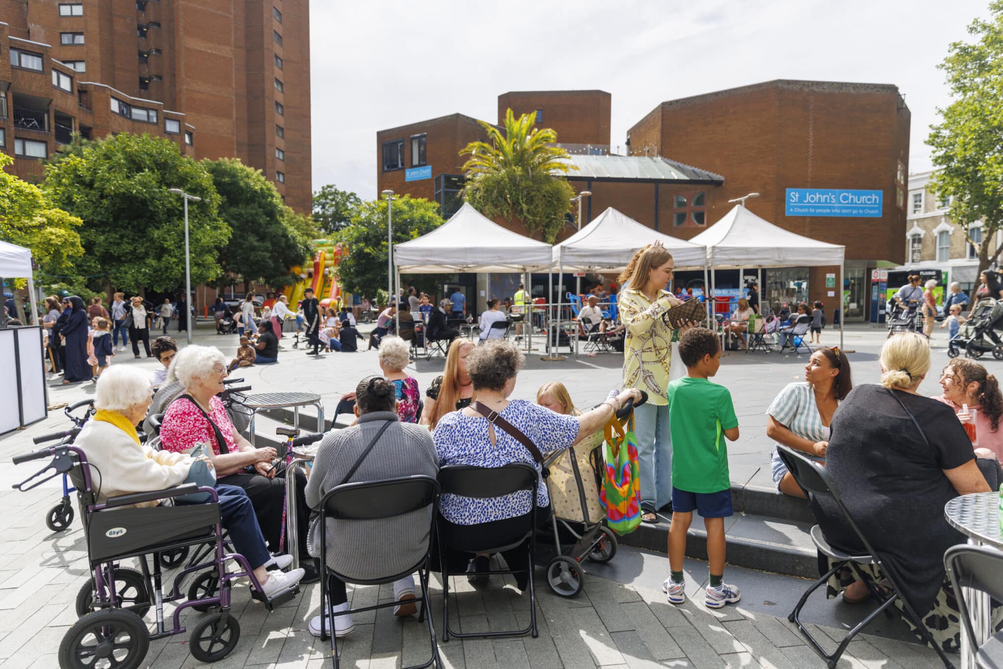 A colourful scene on a sunny day at a community summer event. Children and adults are standing and sitting, chatting to each other. In the back ground there are white marquee tents with more people engaged in activities and behind them tall red brick buildings.