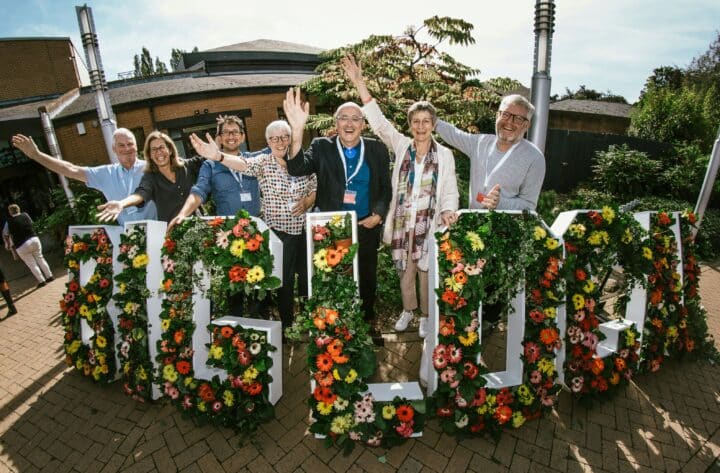 Adults smiling and waving at the camera behind a Big Local flower arrangement