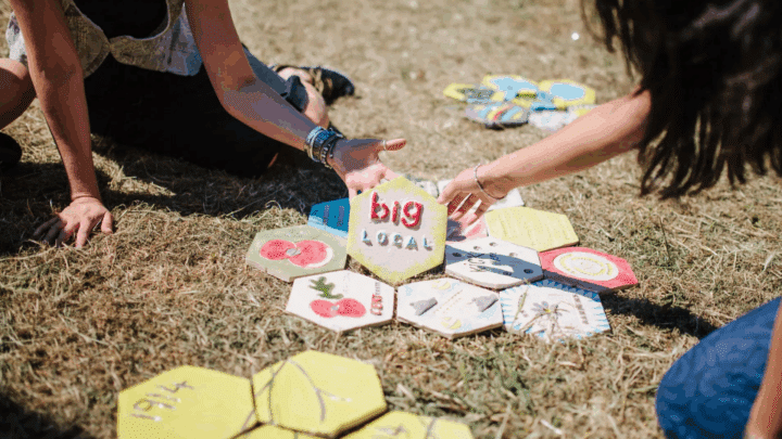 Two people kneel on dry grass, reaching toward a cluster of hand-painted hexagonal tiles arranged like a flower; the center tile reads “Big Local” in red and blue letters. Surrounding tiles show illustrations of fruits, leaves, and patterns in bright colours, with additional tiles scattered nearby.