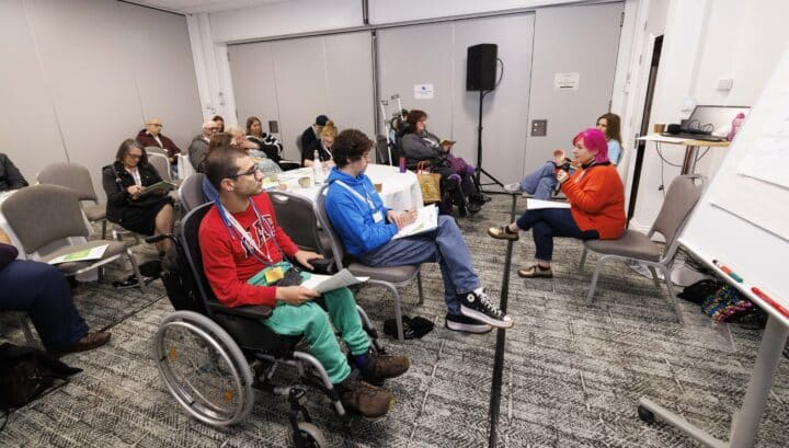 A diverse group of attendees participate in a workshop session. In the foreground, a young man in a red sweatshirt and green trousers uses a wheelchair and holds papers, next to a person in a blue hoodie holding notes. Facing them, a woman with bright pink hair in an orange jumper speaks to the group, with a flipchart beside her. Other attendees are seated on chairs around the room, listening and taking notes.