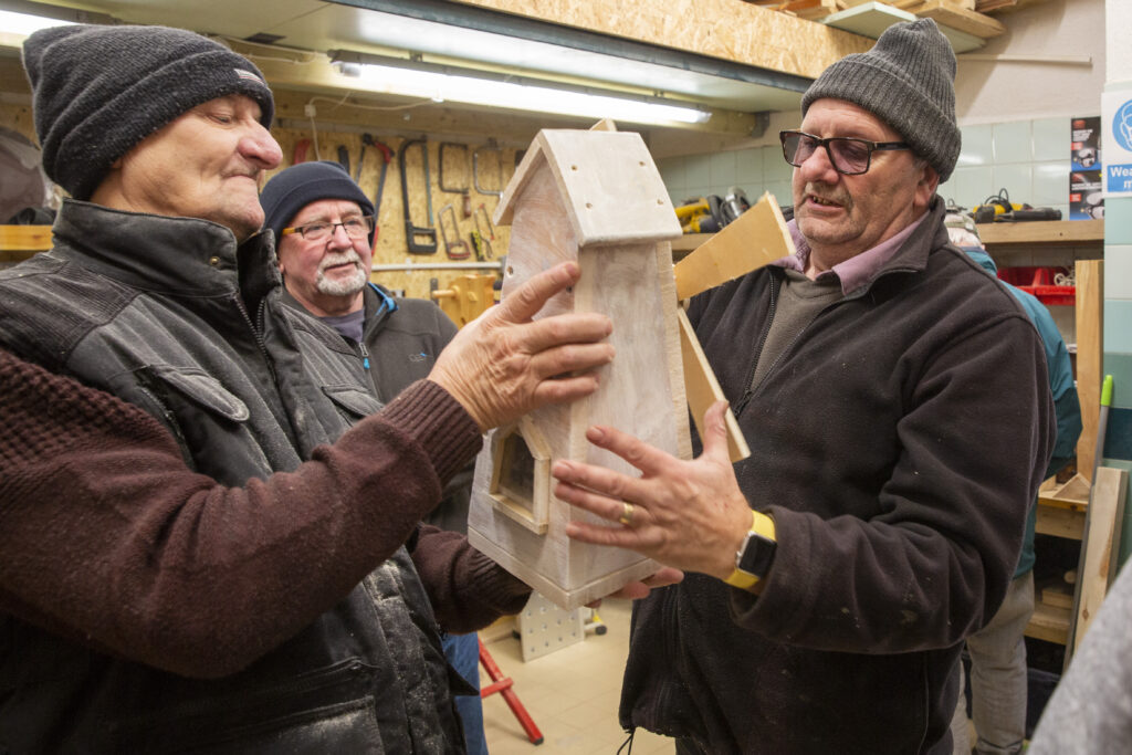 Three older men wearing hats and warm clothing examine a wooden birdhouse inside a workshop, holding it together at chest height; tools hang on the wall behind them and shelves with equipment and materials line the space.