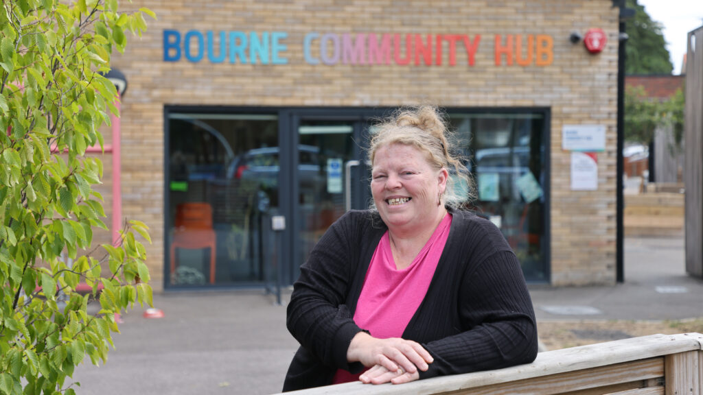 A woman wearing a pink top and black cardigan leans on a wooden fence, smiling. Behind her is a single storey brick building, with a sign above the doors that reads 'Bourne Community Hub'.