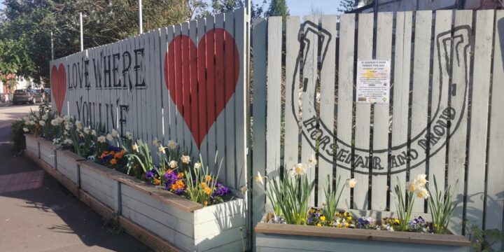 A painted wooden fence decorated with spring flowers in raised planters. The left side reads 