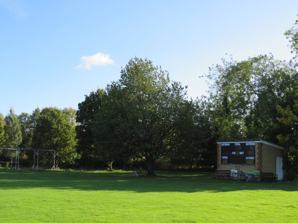 A sunny English village cricket ground with a traditional brick scoreboard and lush green outfield surrounded by trees.