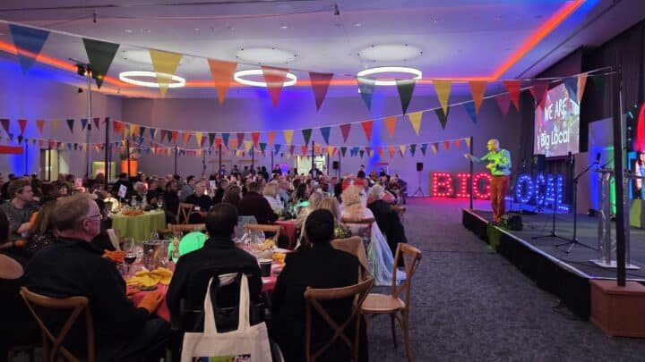 A large indoor celebration event with guests seated at colourful round tables, decorated with bunting flags in red, yellow, green and blue. A speaker stands on a stage to the right beside illuminated marquee letters spelling 'BIG LOCAL' and a projection screen reading 'We Are Big Local
