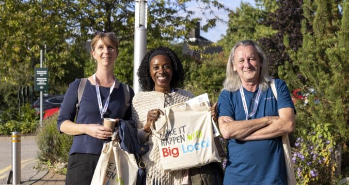 Man and two women standing outside a conference centre smiling at the camera, holding up a bag which says 'Make it happen. Big Local'.