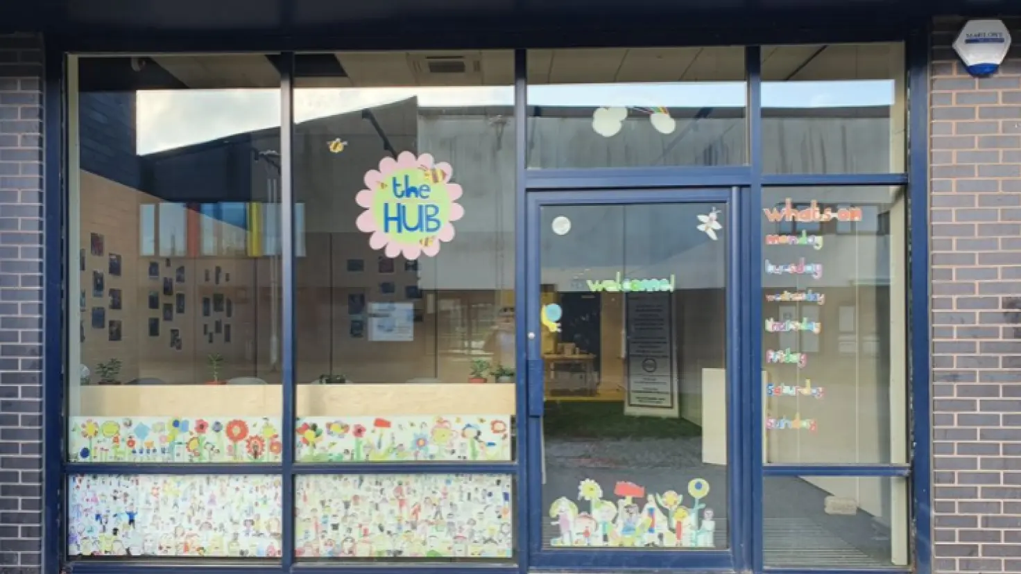 The entrance of a community hub, showing large glass windows decorated with colourful children's artwork and flower designs. A pink flower-shaped sign reads 'the Hub', and the entrance door displays a 'welcome' sign alongside a weekly schedule of activities