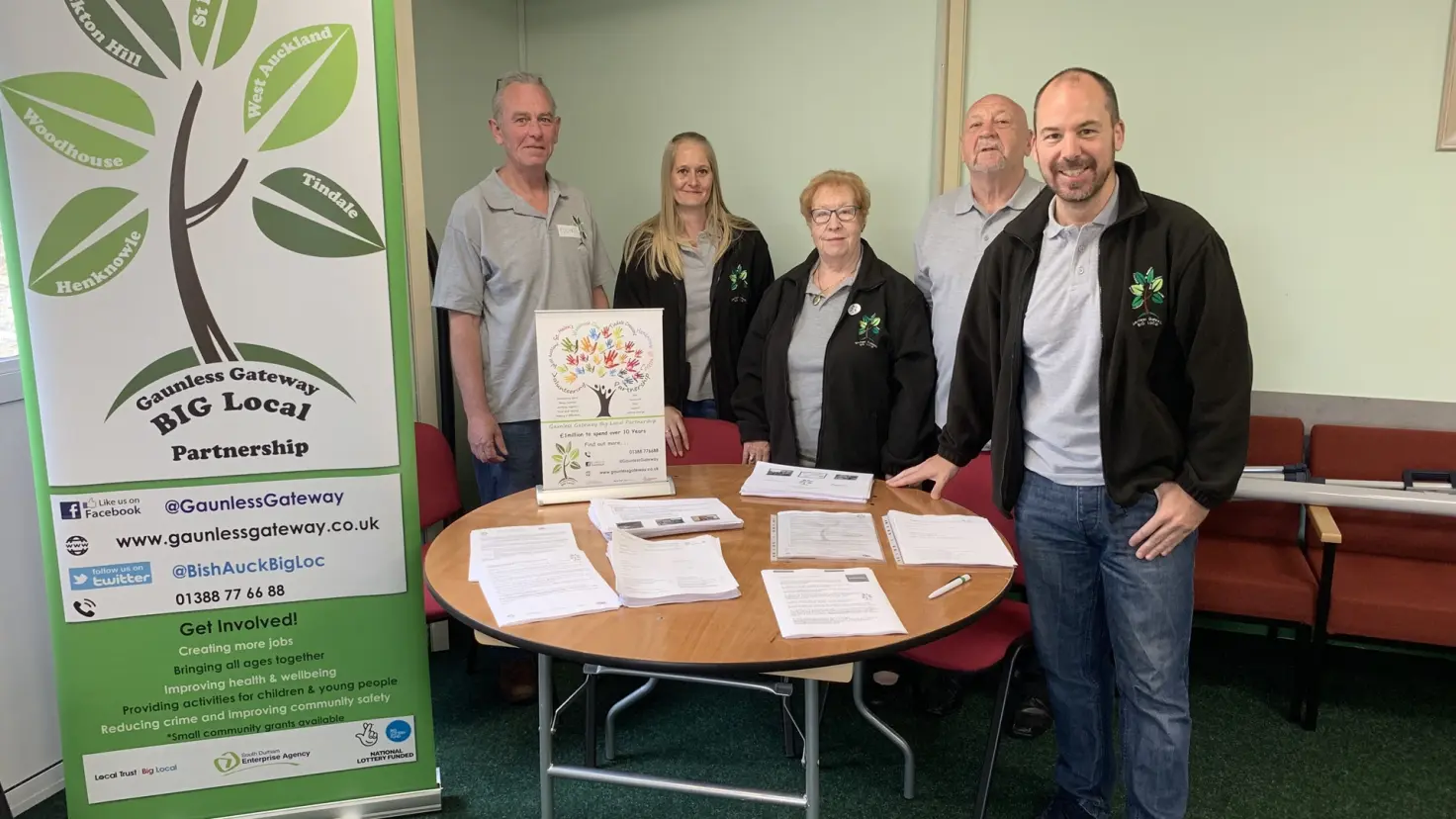 A group of five adults stand behind a table covered with documents, posing beside a banner for the Gaunless Gateway Big Local Partnership.