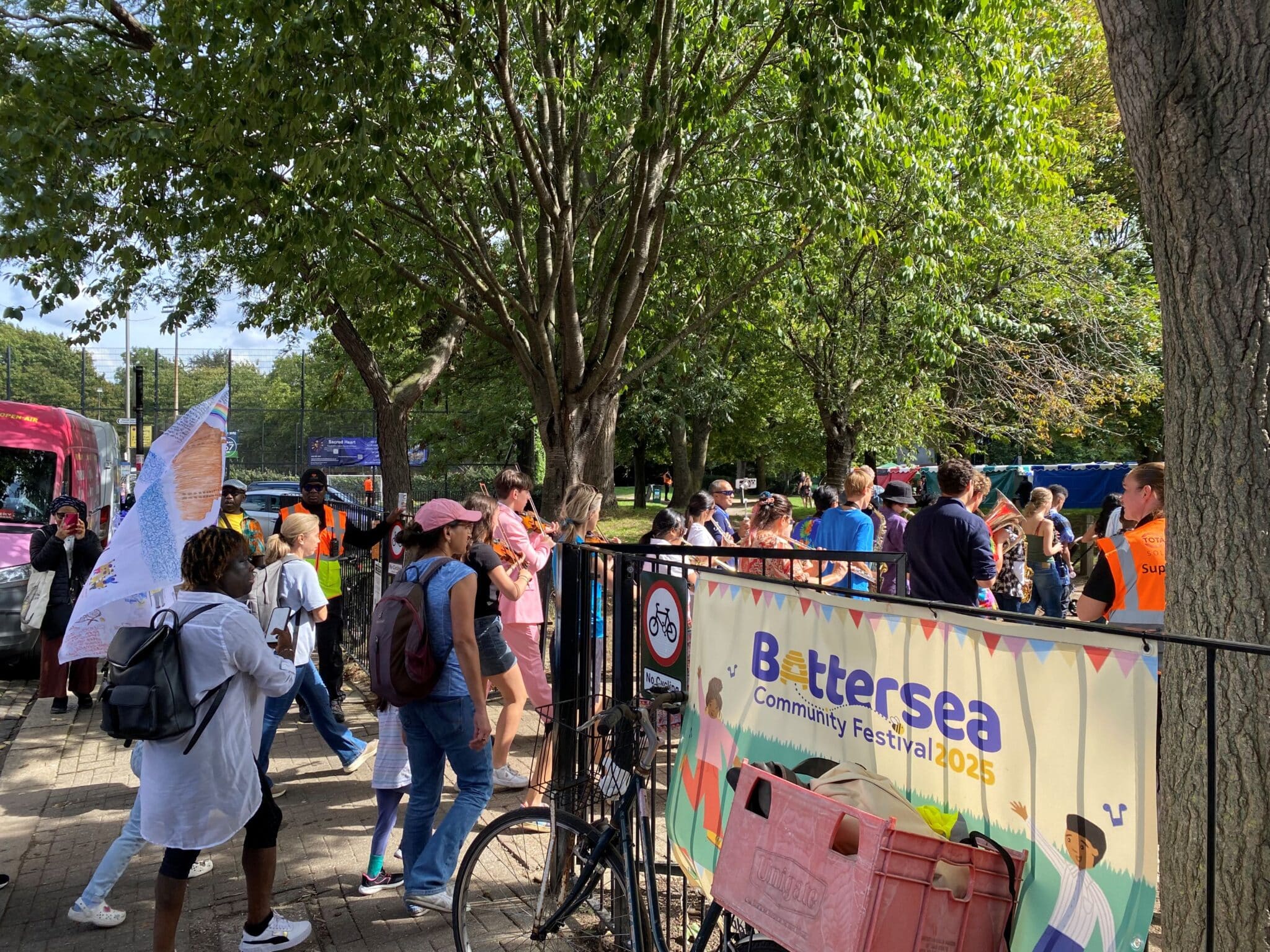A crowd of people gathering at the entrance to the Battersea Community Festival 2025, with a violinist performing in pink and a colourful festival banner on the railings, surrounded by trees on a sunny day