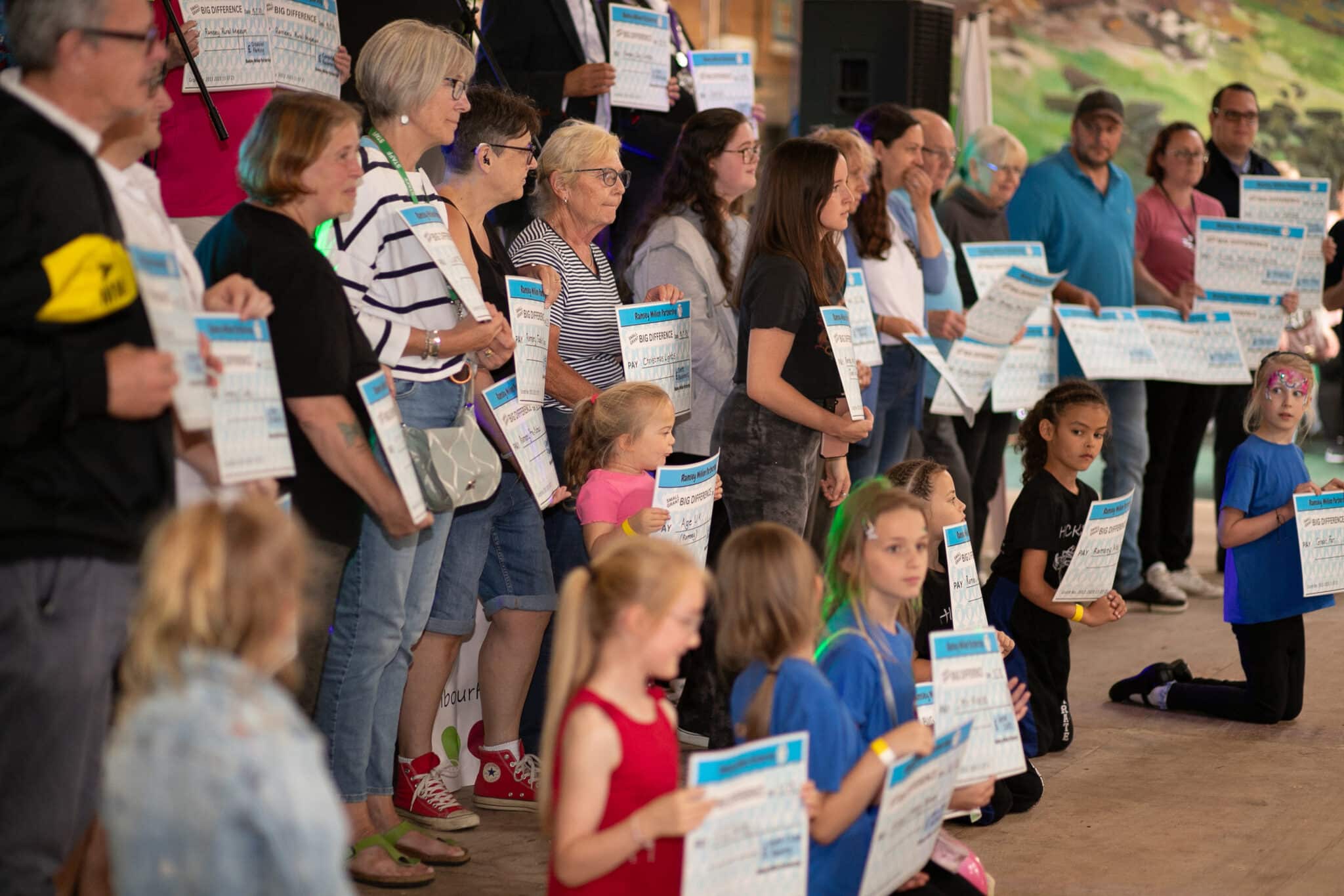A large group of adults and children standing and sitting together at an event, each holding oversized cheques branded with 'Big Difference' at a celebratory community gathering."