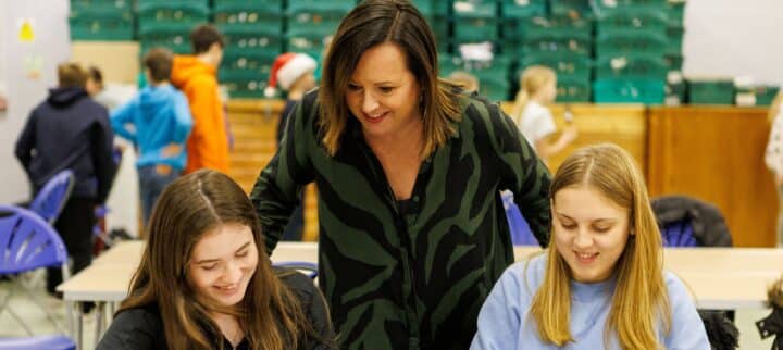 An adult woman leans over two girls sitting at a table as they draw and decorate craft materials with colorful markers. The table is covered with art supplies, and other children can be seen in the background of a busy indoor activity space