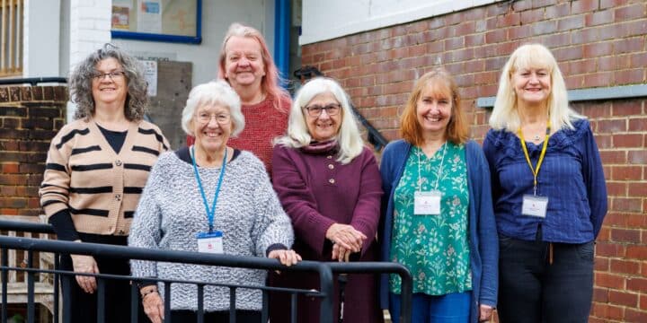 A group of six people smiling and posing together outside a brick building, some wearing lanyards and name badges, gathered near a metal railing.