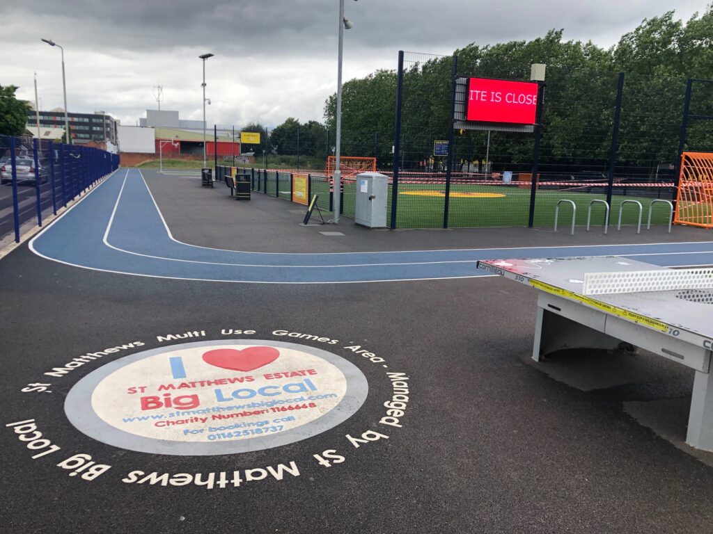 An empty outdoor multi-use games area at St Matthews Estate Big Local, with a blue running track curving around a fenced sports pitch. A red digital sign reads “Site is Closed,” and a table tennis table sits in the foreground beside a large circular ground logo.