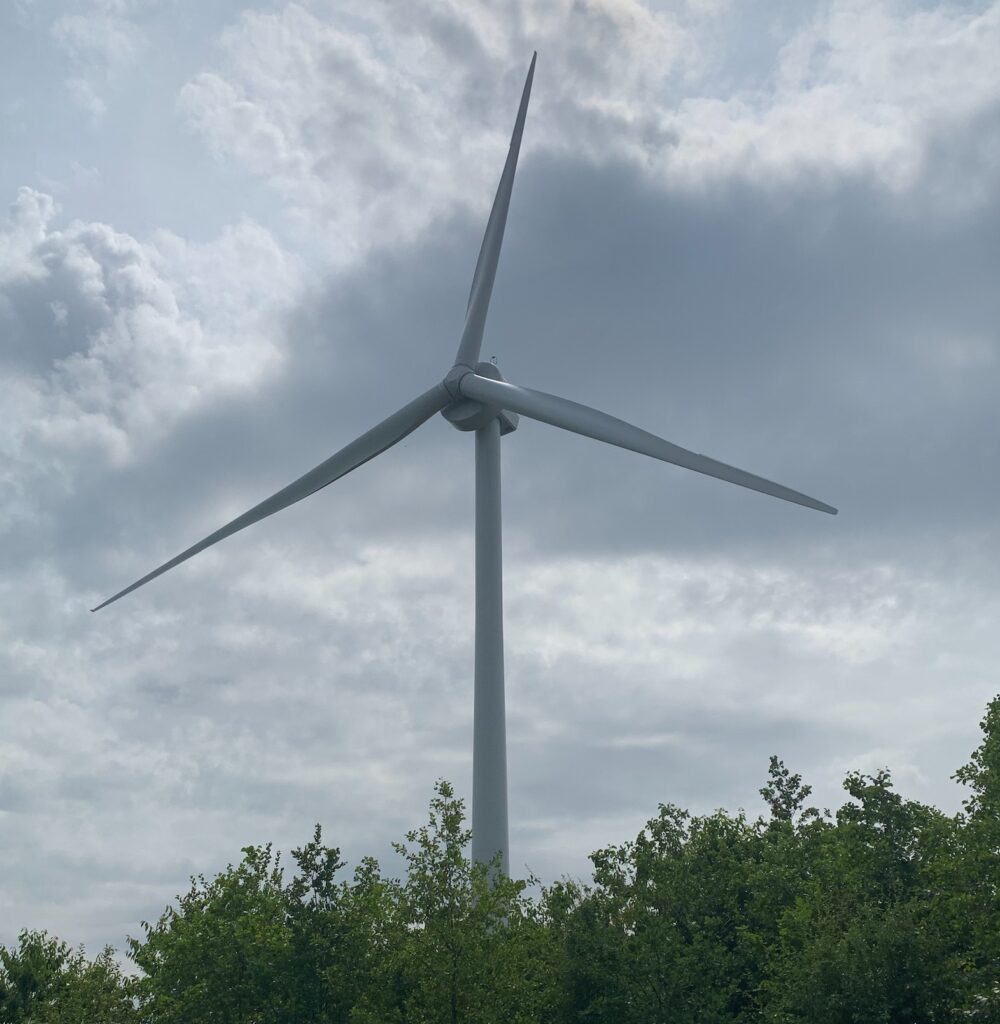 A large white wind turbine rising above green trees against a cloudy sky.