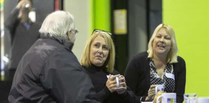 Three people in conversation at an indoor event, holding mugs of tea or coffee, against a bright green wall. Two women with blonde hair face the camera while a third person with white hair is seen from behind.
