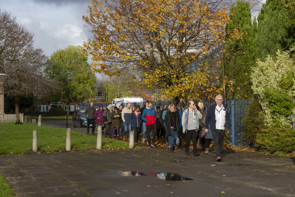 A large group of people walking together along a residential pavement on an autumn day, surrounded by trees with golden leaves, passing blue railings and green grass.