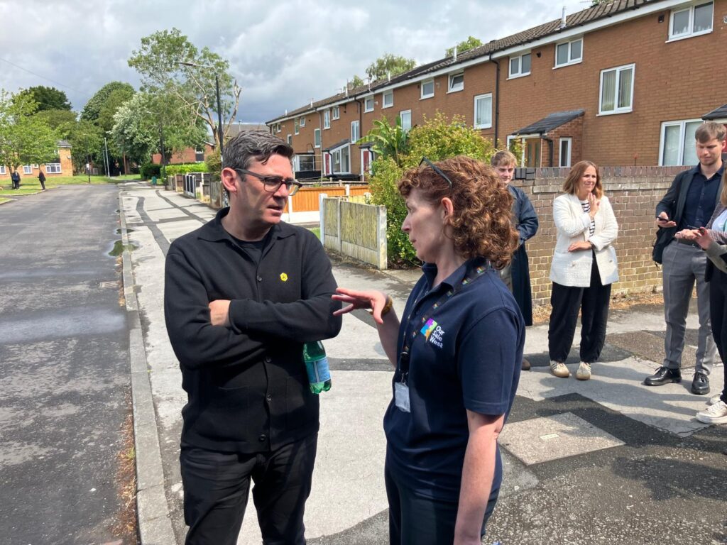 Two people in conversation on a residential street, with a group of people standing in the background. The woman is wearing a navy Our Sale West Big Local polo shirt and lanyard, speaking with a man dressed in black who is holding a water bottle.