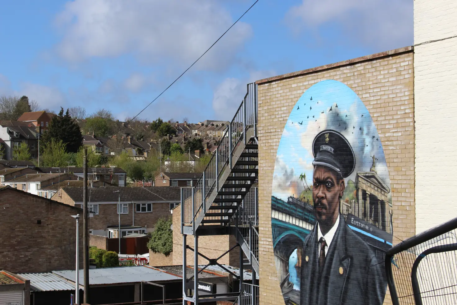 A large mural painted on the side of a brick building depicting a man in a uniform and cap, set against a backdrop of a blue sky and a bridge, with a hillside residential neighbourhood visible in the background.
