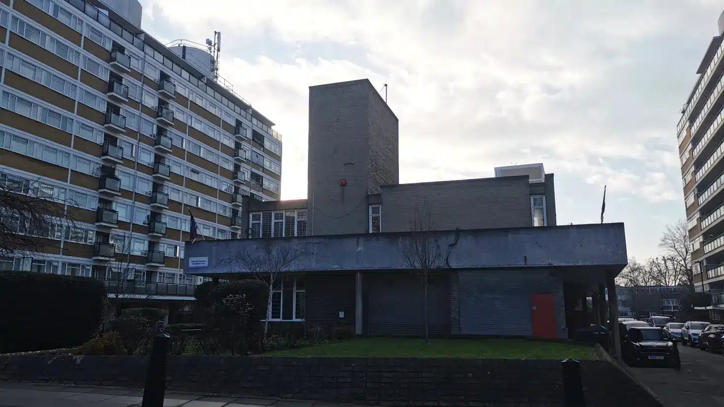 A low-rise brick community building with a distinctive red door sits between two tall residential tower blocks on a cloudy day